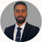 Professional headshot: man in a dark suit, white shirt, navy polka-dot tie, gray circular background.